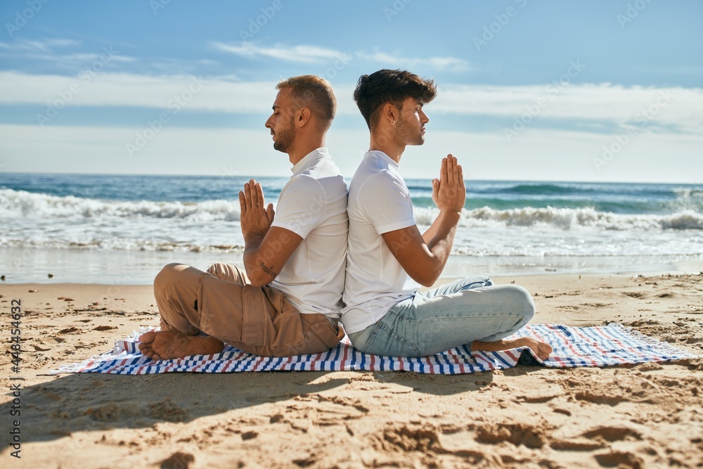 Young gay couple doing yoga sitting at the beach.