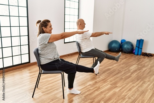 Middle age hispanic couple stretching using chair at sport center.