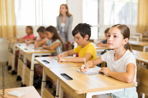 Diligent elementary school student tween girl studying with classmates, making notes of teacher lecture