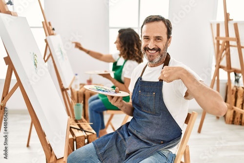 Hispanic middle age man and mature woman at art studio pointing finger to one self smiling happy and proud