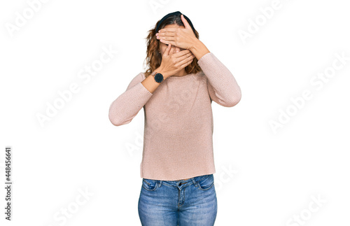 Beautiful young caucasian woman wearing casual sweater covering eyes and mouth with hands, surprised and shocked. hiding emotion