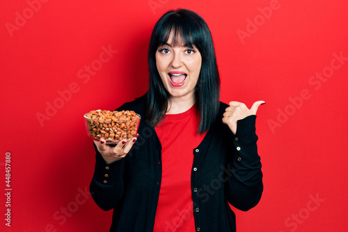 Young hispanic woman holding peanuts pointing thumb up to the side smiling happy with open mouth