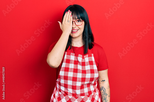 Young hispanic woman wearing cook apron and glasses covering one eye with hand, confident smile on face and surprise emotion. © Krakenimages.com