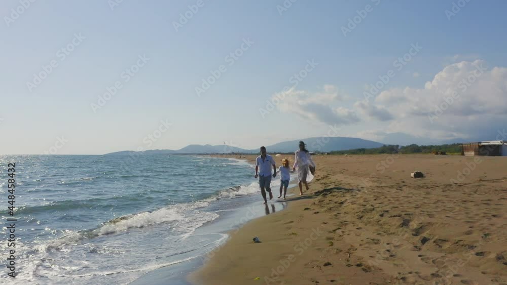Backwards tracking of family running through waves washing sandy beach. People holding hands and enjoying sunny day on coast. Backwards tracking of family running through waves washing sandy beach. People holding hands and enjoying sunny day on coast.