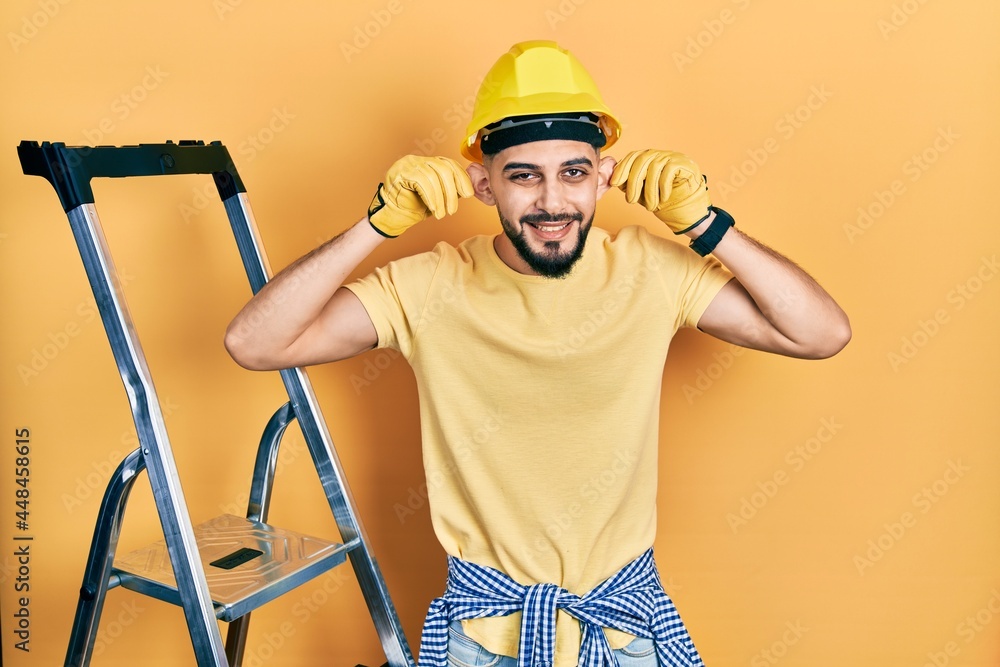 Handsome man with beard by construction stairs wearing hardhat smiling pulling ears with fingers, funny gesture. audition problem Handsome man with beard by construction stairs wearing hardhat smiling pulling ears with fingers, funny gesture. audition problem