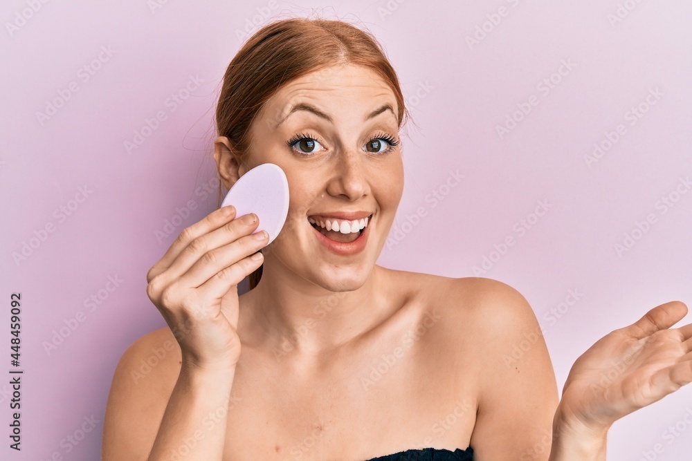 Young irish woman holding makeup sponge celebrating achievement with happy smile and winner expression with raised hand Young irish woman holding makeup sponge celebrating achievement with happy smile and winner expression with raised hand