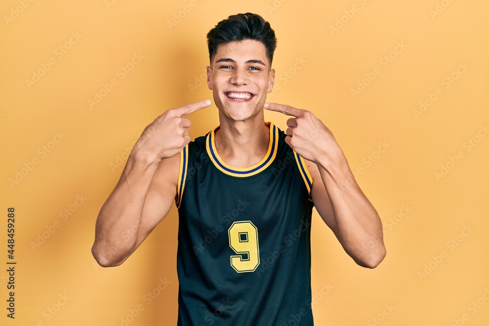 Young hispanic man wearing basketball uniform smiling cheerful showing and pointing with fingers teeth and mouth. dental health concept. Young hispanic man wearing basketball uniform smiling cheerful showing and pointing with fingers teeth and mouth. dental health concept.