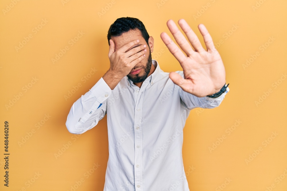 Hispanic man with beard wearing business shirt covering eyes with hands and doing stop gesture with sad and fear expression. embarrassed and negative concept. Hispanic man with beard wearing business shirt covering eyes with hands and doing stop gesture with sad and fear expression. embarrassed and negative concept.