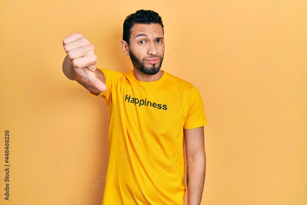 Hispanic man with beard wearing t shirt with happiness word message looking unhappy and angry showing rejection and negative with thumbs down gesture. bad expression. Hispanic man with beard wearing t shirt with happiness word message looking unhappy and angry showing rejection and negative with thumbs down gesture. bad expression.