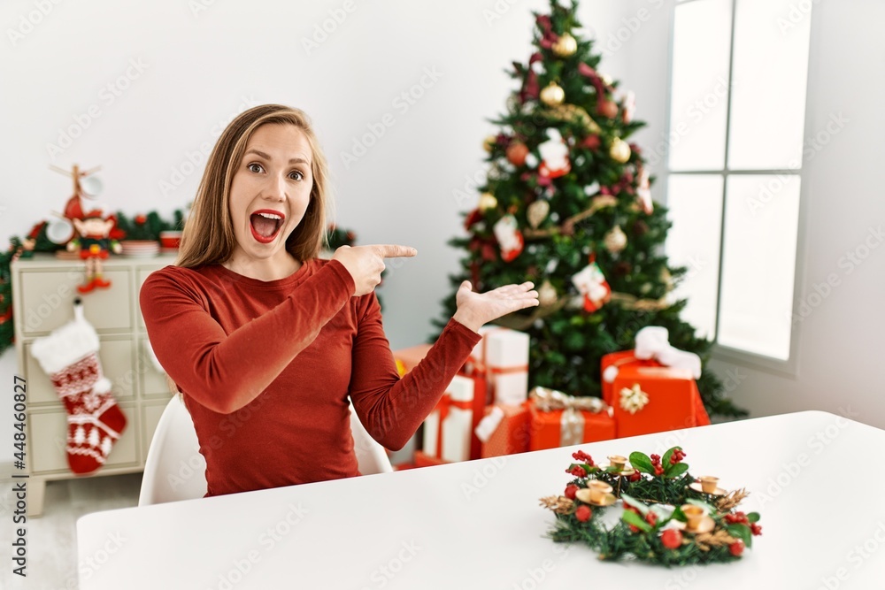 Caucasian young blonde woman sitting on the table by christmas tree amazed and smiling to the camera while presenting with hand and pointing with finger. Caucasian young blonde woman sitting on the table by christmas tree amazed and smiling to the camera while presenting with hand and pointing with finger.