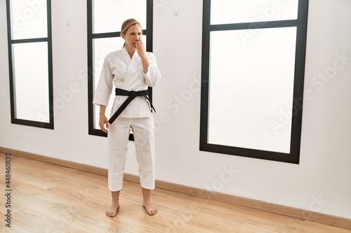 Caucasian young blonde woman wearing karate kimono and black belt looking stressed and nervous with hands on mouth biting nails. anxiety problem.