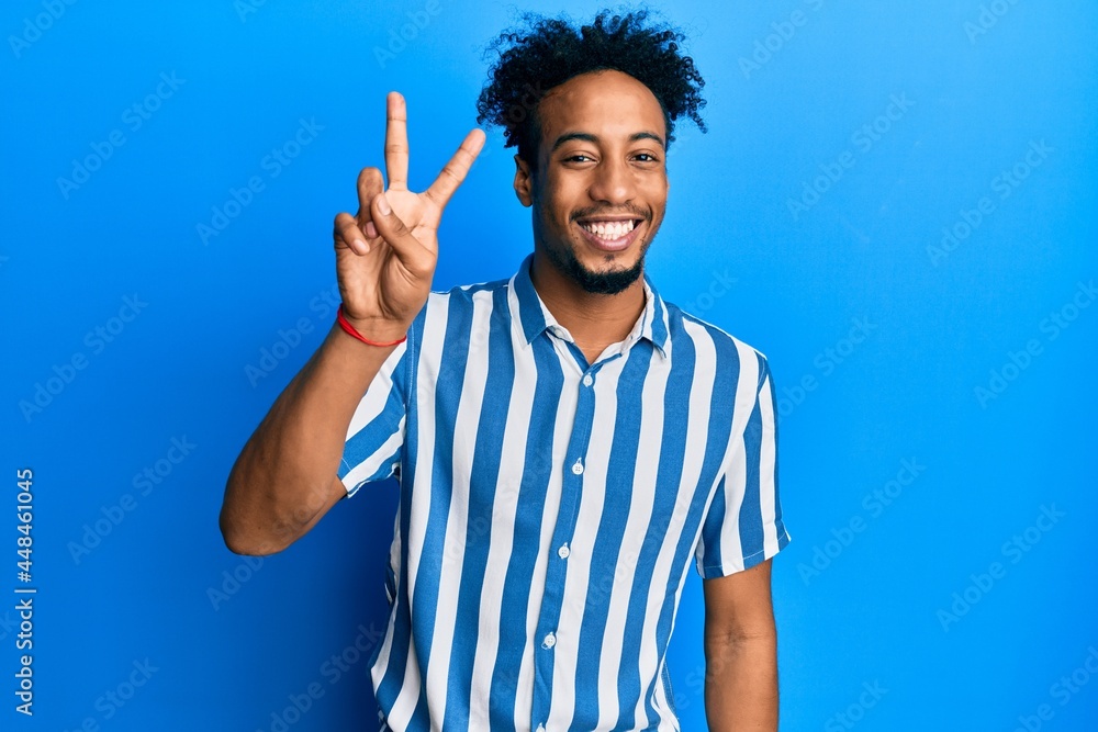 Young african american man with beard wearing casual striped shirt smiling with happy face winking at the camera doing victory sign. number two. Young african american man with beard wearing casual striped shirt smiling with happy face winking at the camera doing victory sign. number two.