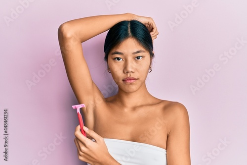 Young chinese woman wearing shower towel using razor relaxed with serious expression on face. simple and natural looking at the camera. photo