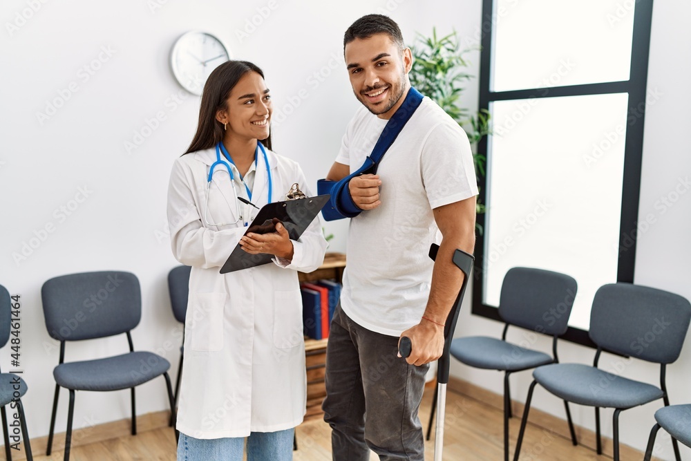 Youg latin injuried man wearing sling and using crutches talking with the doctor at clinic waiting room. Youg latin injuried man wearing sling and using crutches talking with the doctor at clinic waiting room.