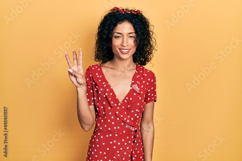 Young latin girl wearing summer dress showing and pointing up with fingers number three while smiling confident and happy.