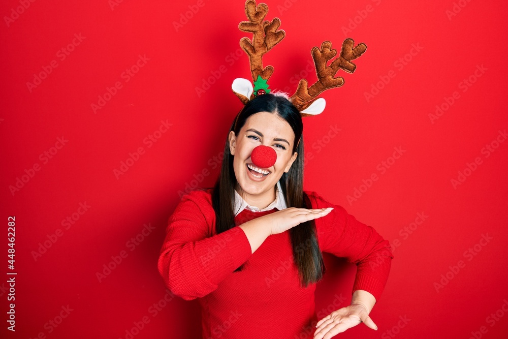 Young hispanic woman wearing deer christmas hat and red nose gesturing with hands showing big and large size sign, measure symbol. smiling looking at the camera. measuring concept. Young hispanic woman wearing deer christmas hat and red nose gesturing with hands showing big and large size sign, measure symbol. smiling looking at the camera. measuring concept.