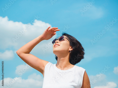 Woman in summertime. Smiling beautiful Asian woman short hair wearing sunglasses and white sleeveless shirt looking up and shading eyes with her hand on blue sky background on sunny day in summer.