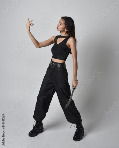 Full length portrait of pretty young asian girl wearing black tank top, utilitarian pants and leather boots. Standing pose holding a knife, isolated against a studio background.