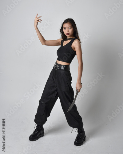 Full length portrait of pretty young asian girl wearing black tank top, utilitarian pants and leather boots. Standing pose holding a knife, isolated against a studio background.