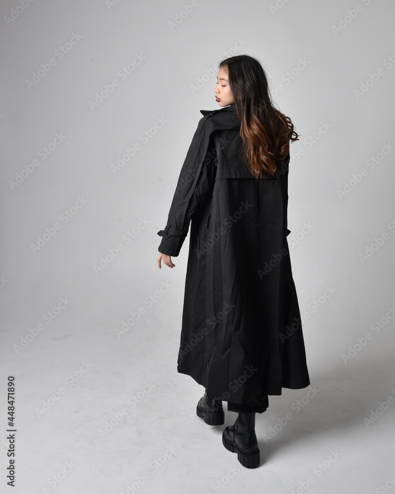 Full length portrait of pretty young asian girl wearing black tank top, utilitarian  pants, leather boots and large dark coat. Standing pose with gestural hand movements,  isolated on  studio backgrou