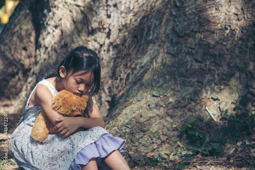 Sad girl hugging teddy bear sitting under tree sadness alone in green park. Lonely girl feeling sad unhappy sit outdoors hug best friend toy. Autism child play teddy bear best friend. Family violence