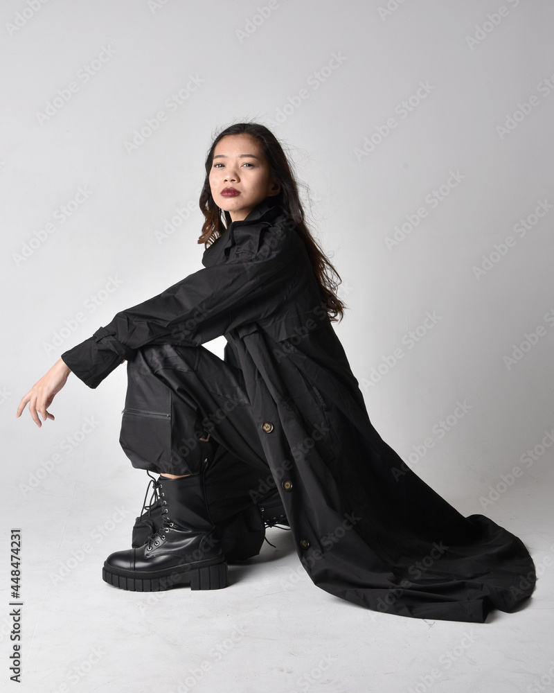 Naklejka premium Full length portrait of pretty young asian girl wearing black tank top, utilitarian pants, leather boots and large dark coat. Sitting pose with gestural hand movements, isolated on studio backgroun