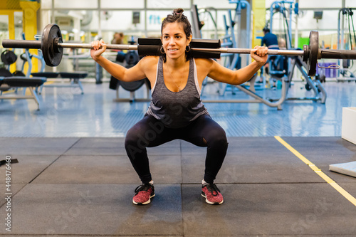 young hispanic latina woman doing barbell squats with weight and a shoulder pad in a gym, smiling. full body