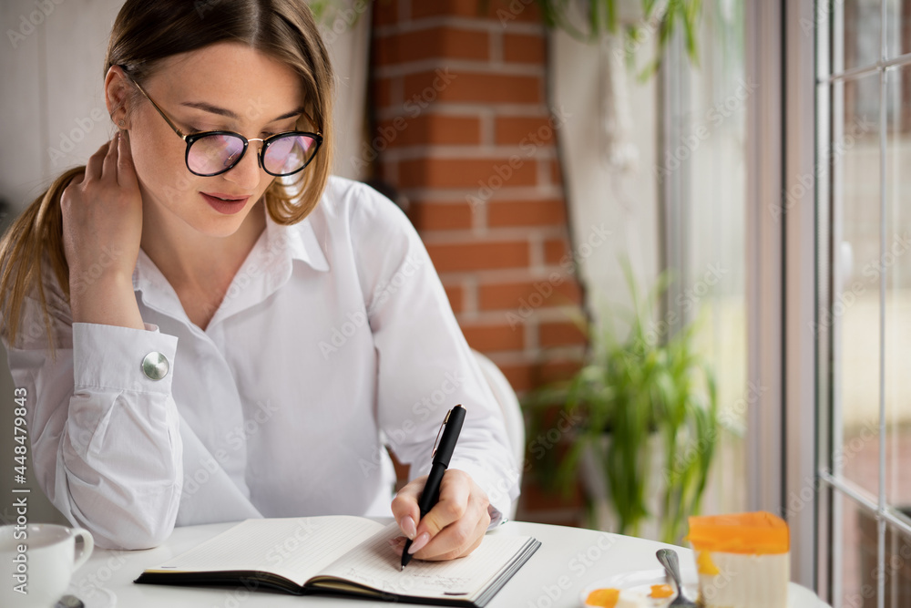 girl writer with glasses sitting in a cafe prepares an article for a newspaper for the publishing house. Happy young woman is writing down business ideas in a notebook. people concept. girl writer with glasses sitting in a cafe prepares an article for a newspaper for the publishing house. Happy young woman is writing down business ideas in a notebook. people concept.