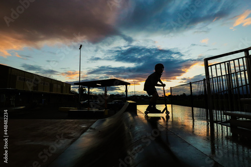 Silhouette image of boy riding scooter at skate park jumping ramp at sunset