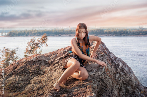 a beautiful slender Indian girl is sitting on a high rock by the river