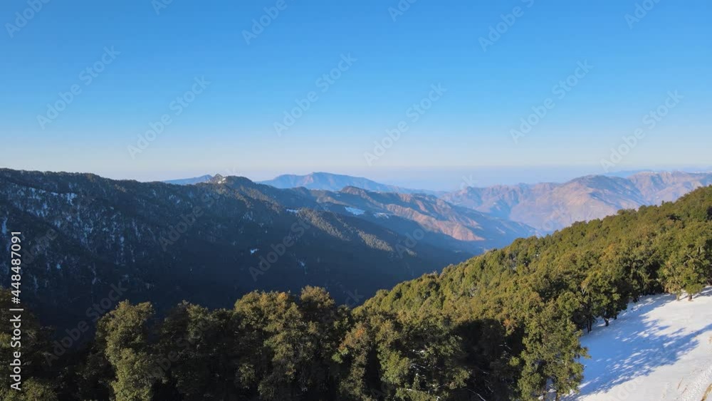 drone moving to the thick vegetation on mountain slope from the snow ground. sunlight on the green trees.blue sky, mountains in background. aerial shot of sunlight and shadow in nature drone moving to the thick vegetation on mountain slope from the snow ground. sunlight on the green trees.blue sky, mountains in background. aerial shot of sunlight and shadow in nature