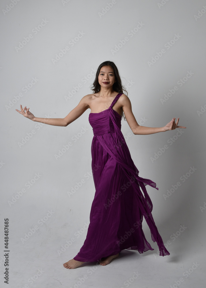 Naklejka premium Full length portrait of pretty brunette asian girl wearing purple flowing gown. Standing, dancing pose on on studio background.
