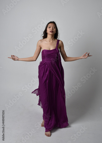 Full length portrait of pretty brunette asian girl wearing purple flowing gown. Standing, dancing pose on on studio background.