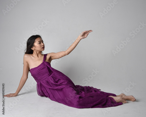 Full length portrait of pretty brunette asian girl wearing purple flowing gown. Sitting pose on on studio background.