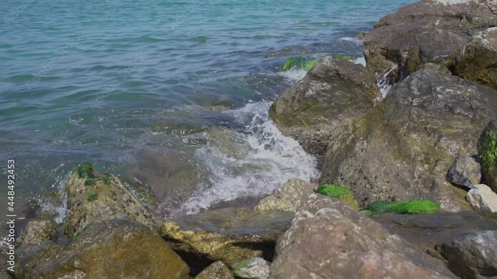 Small blue sea waves roll on grey brown rocks with green moss under bright summer sunlight in tropics slow motion close view Small blue sea waves roll on grey brown rocks with green moss under bright summer sunlight in tropics slow motion close view