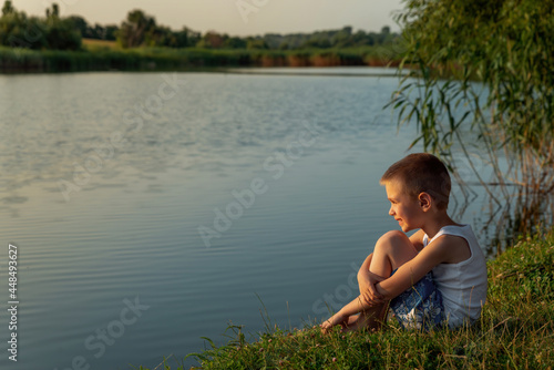 A little happy boy wearing swimming shorts sitting on the shore of a pond at sunset. Concept of freedom and childhood. Child, summer and water. Children's fun in the water. Holidays time