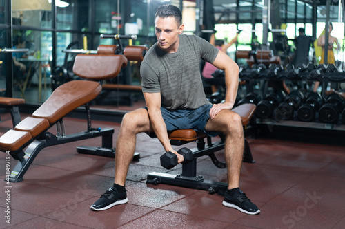Young handsome Caucasian fitness man in sports shirt and shorts, sitting on a fitness bench and exercising with one hand lifting a dumbbells while looking forward in a gym with fitness equipments.
