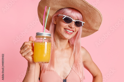 Portrait of happy woman wearing straw hat, swimsuit and sunglasses smiling at camera, drinking tropical juice cocktail, posing isolated over pink studio background. Summer vacation concept