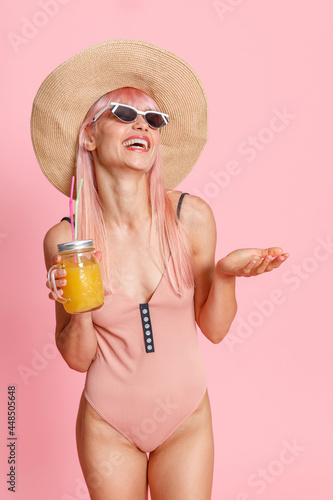 Joyful young woman wearing straw hat, swimsuit and sunglasses holding tropical juice drink, posing isolated over pink studio background. Summer vacation concept