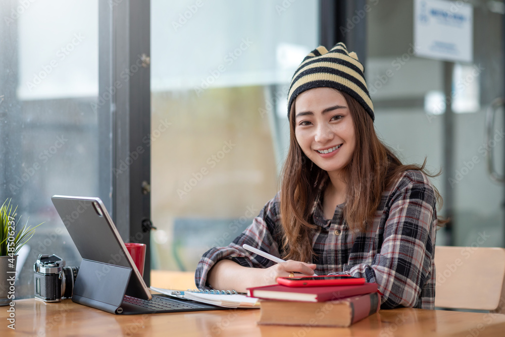 Fototapeta premium Young Asian woman wearing a balaclava sitting at work using a taking note tablet. Looking at the camera.
