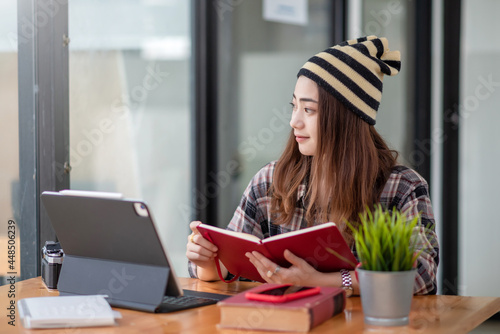 Side view of an Asian woman wearing a balaclava holding a book looking out of the window with a tablet placed at the table.