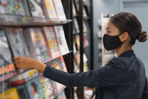 Shopping indoors during coronavirus pandemic. Asian woman wearing face mask while at public library or buying reading magazine at newsstand store in airport. photo