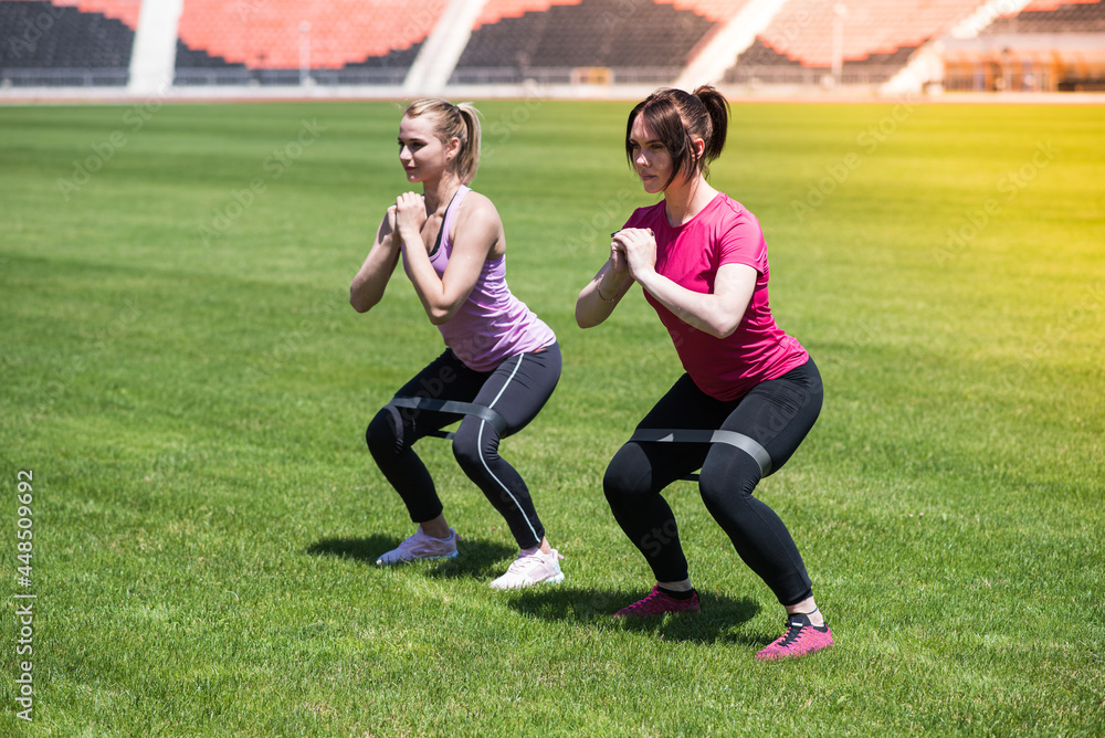 Two women squat on the grass in the stadium. Gymnastics, weight loss, sport