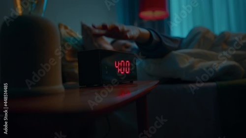 Young sleeping woman wakes up early in morning to disable activated digital alarm clock on bedside table. Doing this she turns away to sleep in covered up in soft blanket. Slow motion cinematic shot photo