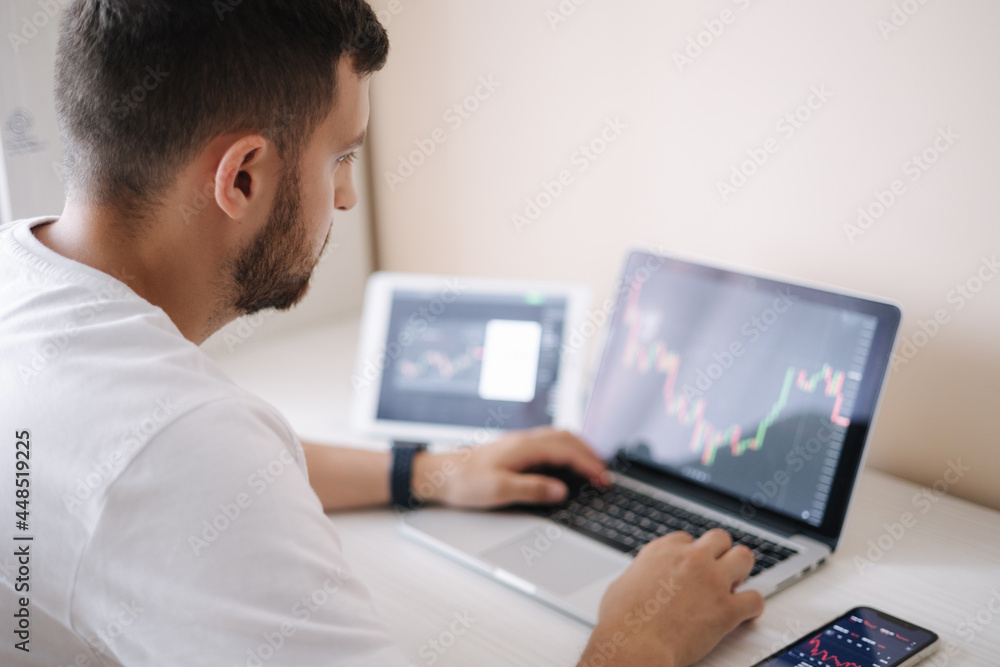 Man looking at finance chart on laptop and tablet, day trader, investing from home. Young man monitors his investments. Ambitious businessman Man looking at finance chart on laptop and tablet, day trader, investing from home. Young man monitors his investments. Ambitious businessman