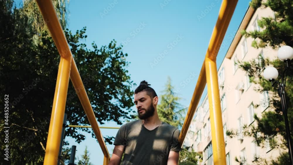 Athletic male is resting on ground near horizontal bar. Caucasian man with beard and dreadlocks stands outside next to parallel bars and takes breath before starting strength training. Athletic male is resting on ground near horizontal bar. Caucasian man with beard and dreadlocks stands outside next to parallel bars and takes breath before starting strength training.