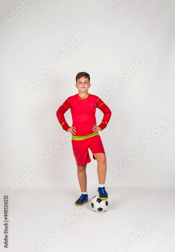 Fototapeta Naklejka Na Ścianę i Meble -  a young football player in a red uniform stands with a soccer ball on a white background with a place for text