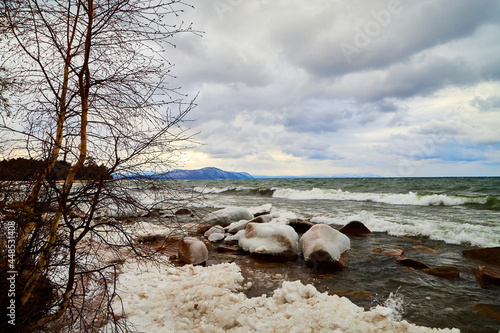 Nature landscape with dramatic sky and clouds, lake with ice, shore with snow and dark trees in cold winter or autumn day or evening