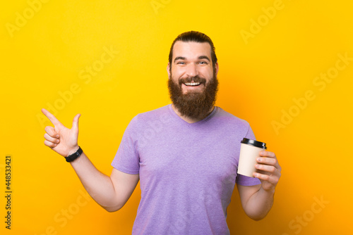 Photo of a happy bearded man holding a cup of hot drink pointing away smiling at the camera