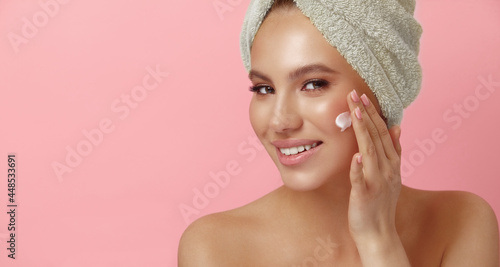 Smiling young woman with clean skin applies cream on face showing jar on pink